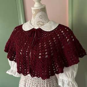 May include: A burgundy crocheted shawl with a scalloped edge and a tie at the neck. The shawl is worn over a white blouse with a Peter Pan collar.