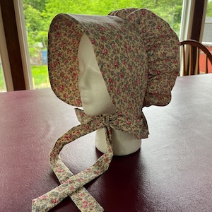 May include: A bonnet featuring a beige fabric base with a dense, repeating pattern of small pink roses and tiny blue flowers. The headwear has a wide, structured brim, a gathered back section, and two long fabric ties that hang down from the chin area.