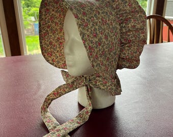 Puede incluir: Un gorro de tela beige con un patrón denso y repetitivo de pequeñas rosas rosadas y diminutas flores azules. La prenda cuenta con un ala ancha y estructurada, una sección trasera fruncida y dos largas cintas de tela que cuelgan bajo la barbilla.