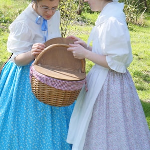May include: A round woven wicker picnic basket with a hinged wooden lid and a red and white chequered fabric lining. The basket features a sturdy arched handle and is held by two individuals wearing long skirts and white blouses.
