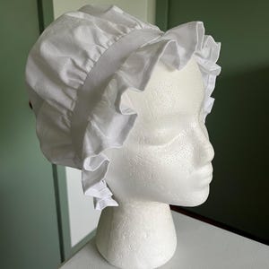 Puede incluir: Un gorro blanco de algodón con volantes, exhibido sobre una cabeza de poliestireno blanco. El gorro tiene una corona fruncida y un borde ancho con volantes. El fondo es verde claro y blanco.
