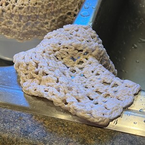 May include: A white crocheted dishcloth with a delicate pattern, sitting on a stainless steel sink.