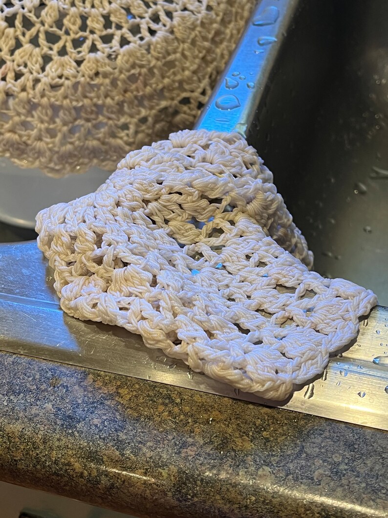 May include: A white crocheted dishcloth with a delicate pattern, sitting on a stainless steel sink.