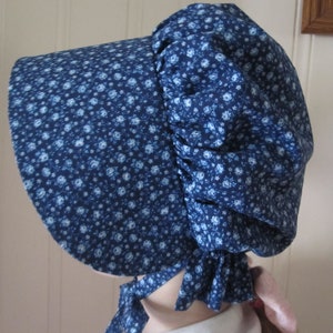 May include: A blue bonnet with a floral pattern. The bonnet features a wide brim and a gathered back, with ties under the chin. The fabric is a dark blue with small, light blue flowers.