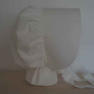 May include: A solid white bonnet featuring a structured, rounded front brim and a gathered, pleated back section. The headwear includes long, flat fabric ties extending from the base, designed to be fastened under the chin for a secure fit.