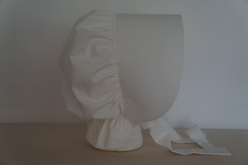 May include: A solid white bonnet featuring a structured, rounded front brim and a gathered, pleated back section. The headwear includes long, flat fabric ties extending from the base, designed to be fastened under the chin for a secure fit.