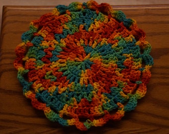 Op de afbeelding: Een handgemaakt, cirkelvormig gehaakt item met een geschulpte rand. Het stuk heeft een levendige reeks kleuren, waaronder oranje, rood, geel, blauwgroen en groen, gerangschikt in een spiraalpatroon. Het item is waarschijnlijk een onderzetter of een trivet.