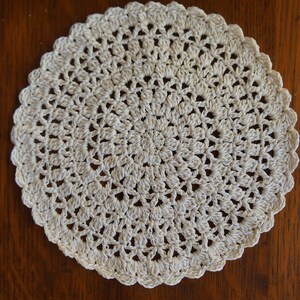 May include: A white crocheted doily with a circular design. The doily is made of delicate, intricate stitches and has a scalloped edge.