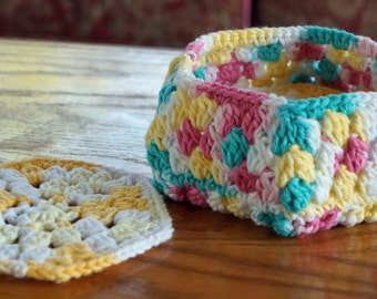 Op de afbeelding: Een gehaakte zeshoekige mand met een roze, gele, witte en turquoise kleurenschema. De mand is leeg en staat op een houten oppervlak.