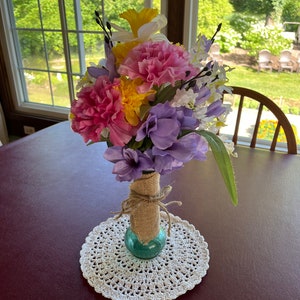 May include: A bouquet of pink, yellow, and purple flowers in a blue glass vase wrapped in burlap. The vase is sitting on a white crocheted doily on a burgundy table.