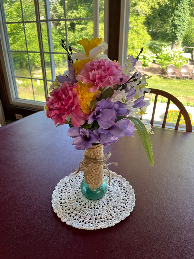 May include: A bouquet of pink, yellow, and purple flowers in a blue glass vase wrapped in burlap. The vase is sitting on a white crocheted doily on a burgundy table.