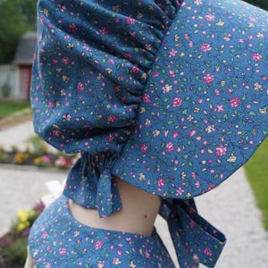 May include: A blue bonnet and matching garment, both adorned with a delicate floral pattern in pink and white. The bonnet features a wide brim and ties, suggesting a vintage or cottagecore aesthetic.