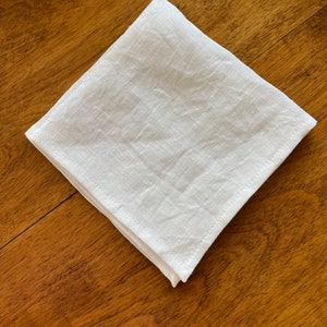 May include: A white linen napkin folded into a square shape, lying on a wooden surface.