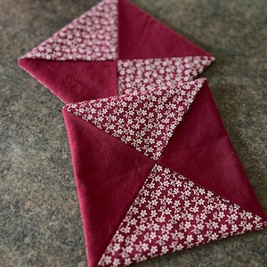 Könnte beinhalten: Zwei quadratische weinrote Stoffuntersetzer mit weißen floral gemusterten Ecken. Die Untersetzer bestehen aus einem weichen Material und sind auf einer dunkelgrauen Oberfläche angeordnet. Die Untersetzer sind handgefertigt.