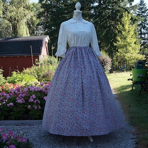 Puede incluir: Un vestido largo con cuerpo blanco, cuello redondo y mangas tres cuartos, combinado con una falda larga fruncida de tela azul claro con un patrón denso y repetitivo de pequeñas flores rojas y rosas en toda la superficie.