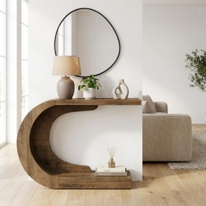Peut inclure: Une console en bois en forme de C avec une finition en grain de bois foncé. La table présente une surface supérieure plate et une étagère de base inférieure. Sur le dessus se trouvent une lampe beige, une petite plante verte et une sculpture.