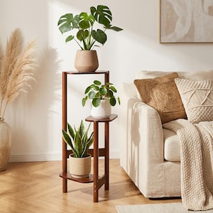 Op de afbeelding: Een drielaagse, donkerbruine houten plantenstandaard met drie potplanten. De planten zijn onder meer een Monstera, een Pilea en een slangengras. De standaard staat naast een beige bank met decoratieve kussens.