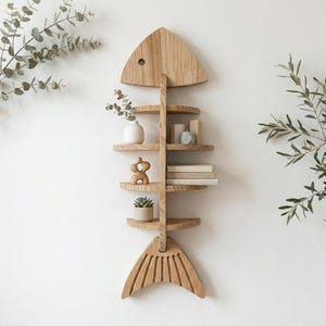 Op de afbeelding: Een houten wandplank in de vorm van een vis, met meerdere ronde planken. De plank is gemaakt van lichtgekleurd hout en is versierd met kleine planten, boeken en decoratieve objecten. Het ontwerp lijkt op een vissenskelet.