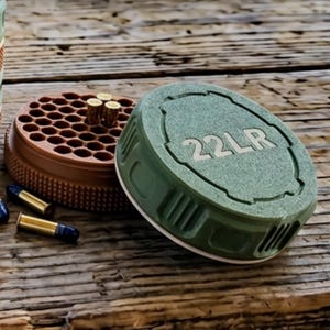 May include: A green and brown ammunition container with the text "22LR" on the lid. The container is open, revealing a brown interior with multiple holes. Several bullets are scattered around the container on a wooden surface.