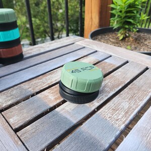 Puede incluir: Una tapa circular de color verde salvia con el texto 45 ACP impreso en negro y amarillo, que presenta un agarre lateral texturizado y una base negra. Una pila de tapas similares en verde, verde azulado y marr&oacute;n es visible al fondo.