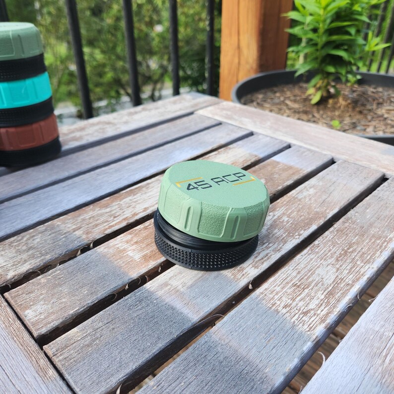 Puede incluir: Una tapa circular de color verde salvia con el texto 45 ACP impreso en negro y amarillo, que presenta un agarre lateral texturizado y una base negra. Una pila de tapas similares en verde, verde azulado y marr&oacute;n es visible al fondo.