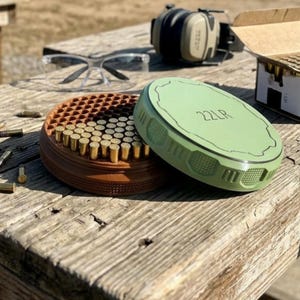 May include: A brown and green ammunition storage container with the text "22LR" on the lid. The container is filled with brass-colored bullets. Safety glasses and ear protection are visible in the background.