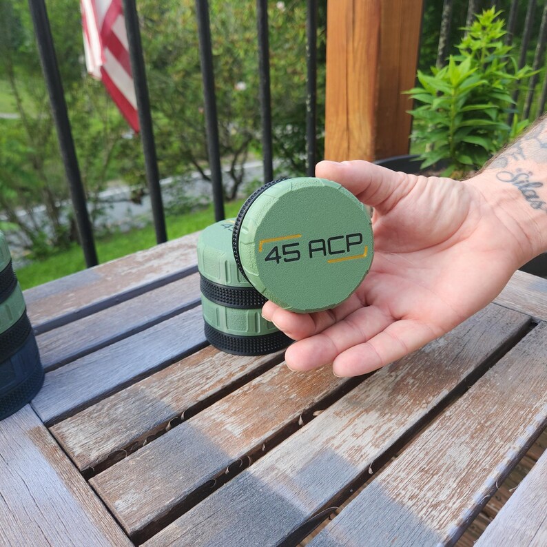 Puede incluir: Una tapa de recipiente redonda de color verde oliva con el texto 45 ACP en letras negras y finas l&iacute;neas horizontales amarillas a ambos lados. La tapa tiene una banda de agarre de goma negra texturizada alrededor de su circunferencia.
