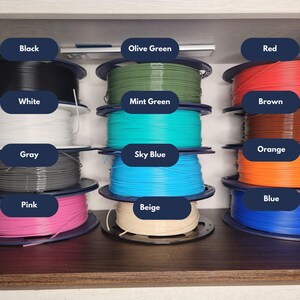 Puede incluir: Una colecci&oacute;n de bobinas de filamento para impresora 3D dispuestas en tres pilas verticales. Los colores incluyen negro, blanco, gris, rosa, verde oliva, verde menta, azul cielo, beige, rojo, marr&oacute;n, naranja y azul. Cada bobina tiene su etiqueta.
