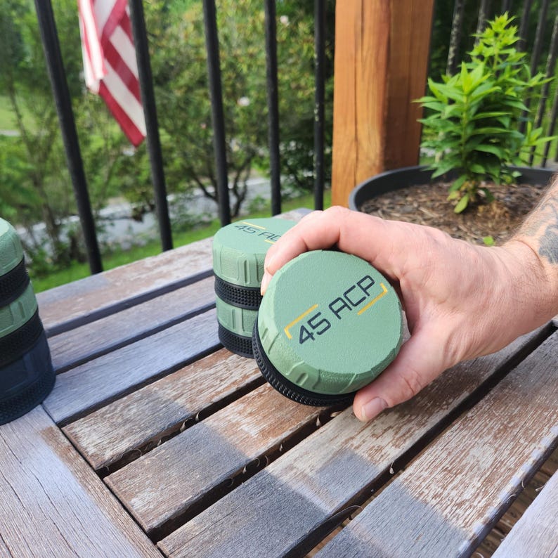 Puede incluir: Un recipiente redondo de color verde oliva con una base negra texturizada y una tapa negra con el texto 45 ACP en una fuente sans-serif en negrita. La tapa tiene dos l&iacute;neas amarillas horizontales y los lados presentan un agarre estriado.