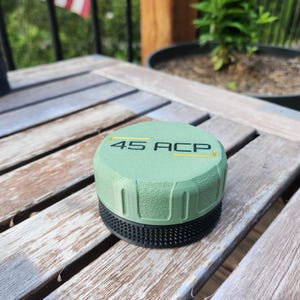 Puede incluir: Una tapa cil&iacute;ndrica de color verde salvia con un lateral texturizado y una base negra. La superficie superior presenta el texto 45 ACP en letras negras, flanqueado por dos l&iacute;neas horizontales amarillas. El objeto tiene un dise&ntilde;o estriado.