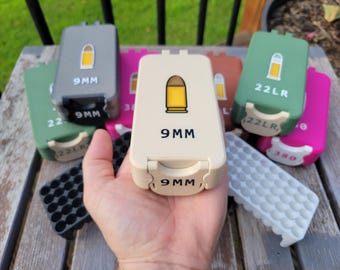May include: A collection of rectangular plastic ammunition storage cases in various colors including cream, grey, green, pink, and brown. Each case features a graphic of a bullet and text indicating the caliber, such as 9MM, 22LR, or 380.