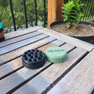 Puede incluir: Un molinillo circular de dos piezas con base negra que presenta un agarre texturizado y una serie de orificios internos. La tapa es de color verde oliva con el texto 45 ACP impreso en negro en la parte superior y un borde estriado.