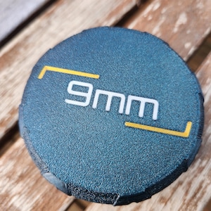 May include: A circular, dark blue, textured object featuring the white text 9mm centered on the surface. The text is framed by two yellow L-shaped corner brackets, one positioned at the top left and one at the bottom right of the lettering.