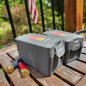 May include: Two gray 12GA ammunition boxes with red and yellow stripes on top. The boxes are on a wooden surface with two shotgun shells in front. The text "12GA" is printed on the boxes.