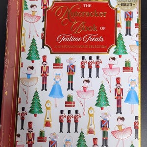 Può includere: Un libro decorativo intitolato "The Nutcracker Book of Teatime Treats" con copertina rossa e dettagli dorati. La copertina presenta illustrazioni di ballerine, schiaccianoci, alberi di Natale e orologi. Contiene biscotti scozzesi.