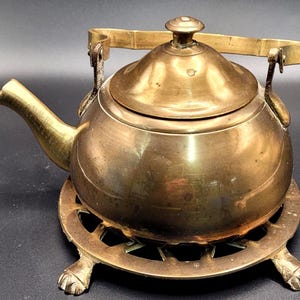 Könnte beinhalten: Eine Vintage-Teekanne aus Messing mit Deckel und Griff. Die Teekanne steht auf einem passenden Messing-Trivet mit dekorativen Füßen. Der Ausguss ist gebogen und der Griff ist gewölbt. Das Gesamtdesign ist klassisch und elegant.