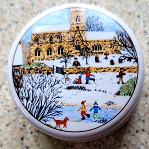 Op de afbeelding: Een ronde, witte sieradendoosdeksel met een wintertafereel. Het toont een kerk, kinderen die in de sneeuw spelen en een rode hond. Het kunstwerk gebruikt een reeks kleuren, waaronder blauw, bruin en rood.