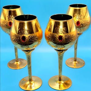 May include: Four ornate gold-colored wine glasses with a red jewel accent. The glasses have a decorative design on the bowl and a twisted stem. The rims are dark green. The glasses are on a blue background.