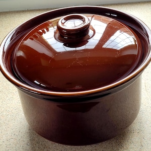 May include: A round, deep, dark brown ceramic cooking pot with a matching lid. The pot features a smooth, glossy, reflective glaze finish. The lid has a small, centred, circular knob handle on top, and the pot has a wide, open rim design.