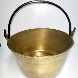 May include: A brass bucket with a smooth, polished finish and a wide, open top. The container features a dark, curved metal handle attached to two side brackets, allowing for easy movement. The interior and exterior surfaces are metallic gold.