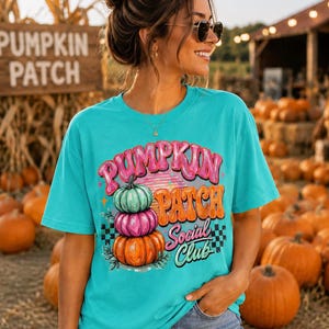 Puede incluir: Una camiseta de color turquesa brillante con un estampado gráfico de tres calabazas apiladas en verde azulado, rosa y naranja. El texto dice PUMPKIN PATCH SOCIAL CLUB en letras estilizadas rosas y naranjas, acentuado por pequeñas estrellas y un patrón de tablero de ajedrez en blanco y negro.