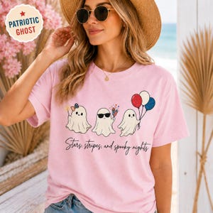 May include: Pink t-shirt featuring three cartoon ghosts with patriotic accessories. One ghost has a bow, another sunglasses, and the third holds balloons. The text "Stars, stripes, and spooky nights" is below the ghosts.