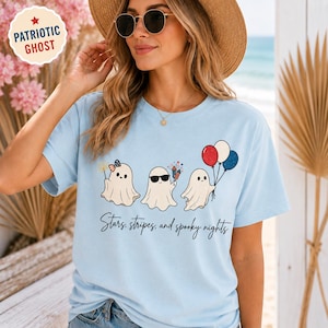 May include: Light blue t-shirt featuring three cartoon ghosts with patriotic accessories. One ghost wears a bow, another sunglasses, and the third holds balloons. The text "Stars, stripes, and spooky nights" is below the ghosts.