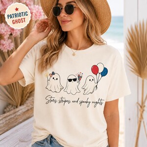 May include: Cream-colored t-shirt featuring three cartoon ghosts with patriotic accessories. One ghost has a bow, another sunglasses, and the third holds red, white, and blue balloons. The text "Stars, stripes, and spooky nights" is below the ghosts.