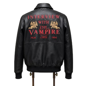 May include: Black leather bomber jacket with a collar and ribbed cuffs. The back features red and gold embroidered text that reads "Interview with the Vampire" with "Film Crew 1994" below, and two angel figures.