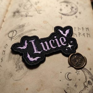 May include: A black embroidered patch with a scalloped edge featuring the name Lucie in light purple gothic-style lettering. The design includes small purple bat silhouettes and white star accents. A circular bronze wax seal is placed nearby.