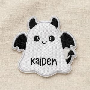 May include: An embroidered iron-on patch shaped like a white ghost with black bat wings, black horns, and a black pointed tail. The ghost has a smiling face with two round black eyes and the name Kaiden stitched in black thread across the center.