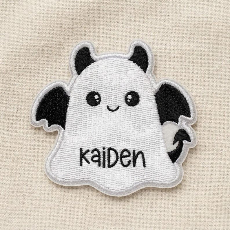 May include: An embroidered iron-on patch shaped like a white ghost with black bat wings, black horns, and a black pointed tail. The ghost has a smiling face with two round black eyes and the name Kaiden stitched in black thread across the centre.