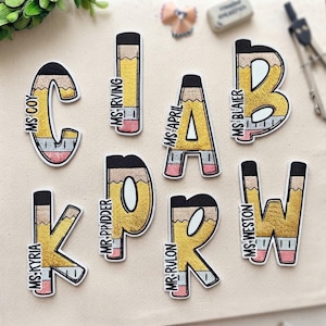 May include: A collection of seven embroidered patches shaped like letters of the alphabet, each designed with a yellow pencil pattern featuring a black tip, a silver ferrule, and a pink eraser. Each patch includes a name printed in black text.