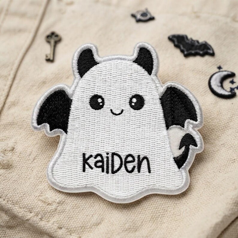 May include: An embroidered patch shaped like a white ghost with black bat wings, black ears, and a black tail. The ghost has a smiling face with two round black eyes and the name "kaiden" stitched in black thread across the lower centre area.